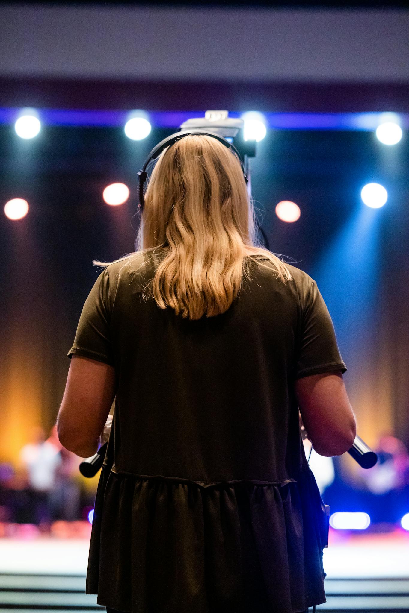 A female videographer with headphones captures an event indoors, focused on a stage.